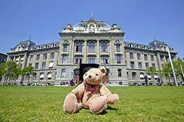 Anlass/Kinderuni/Teddy mit Doktorhut sitzt auf Gras vor dem Hauptgebäude/Sommer